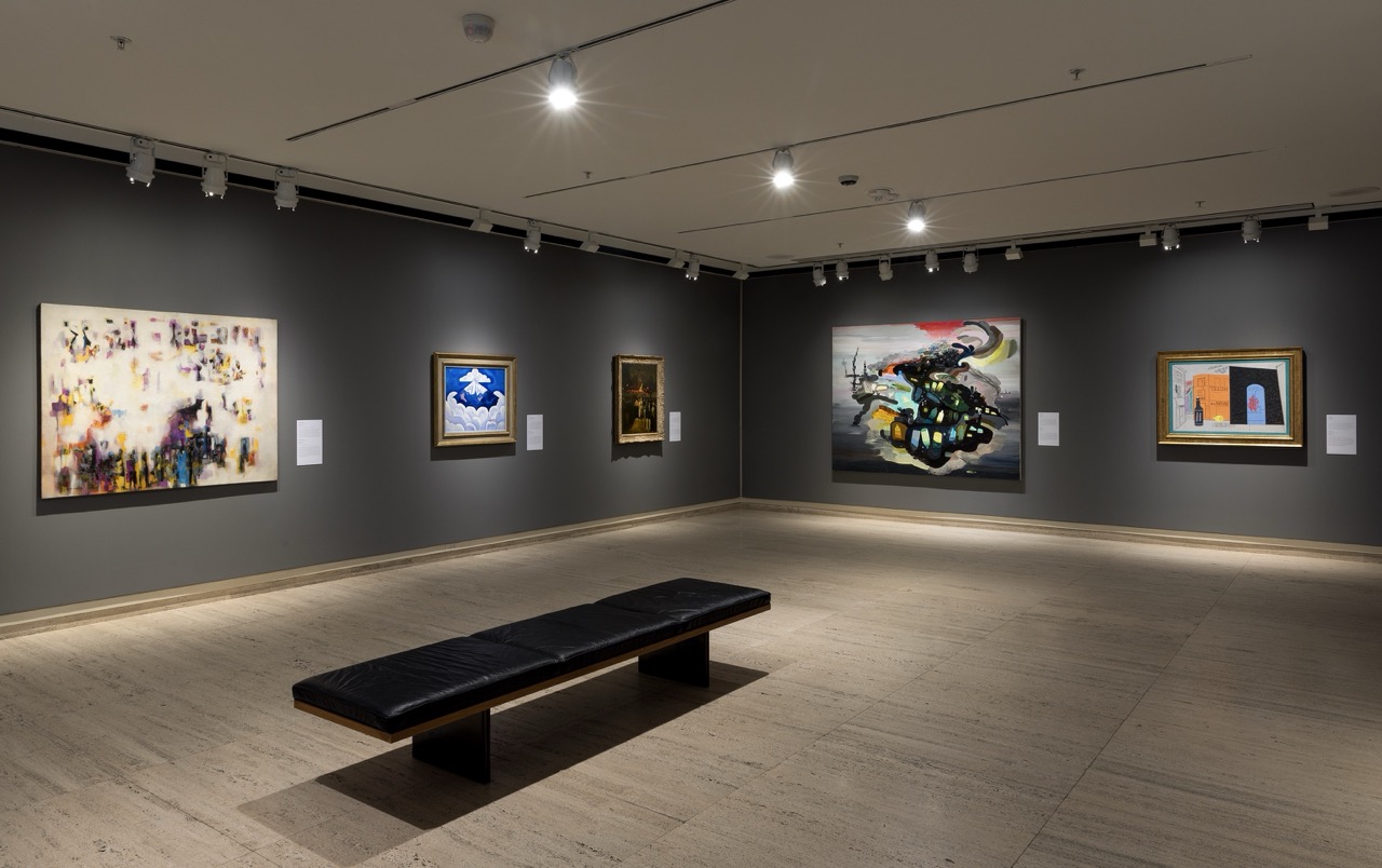 Art gallery with modern and abstract paintings on dark walls, lit by ceiling lights, with a black bench.