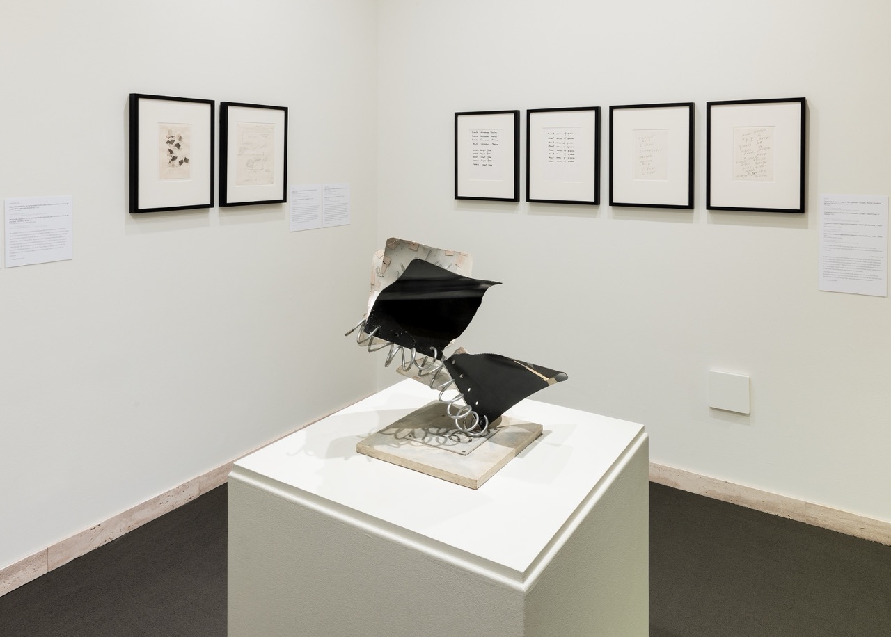 Sculpture on pedestal in gallery with framed artworks on white walls.